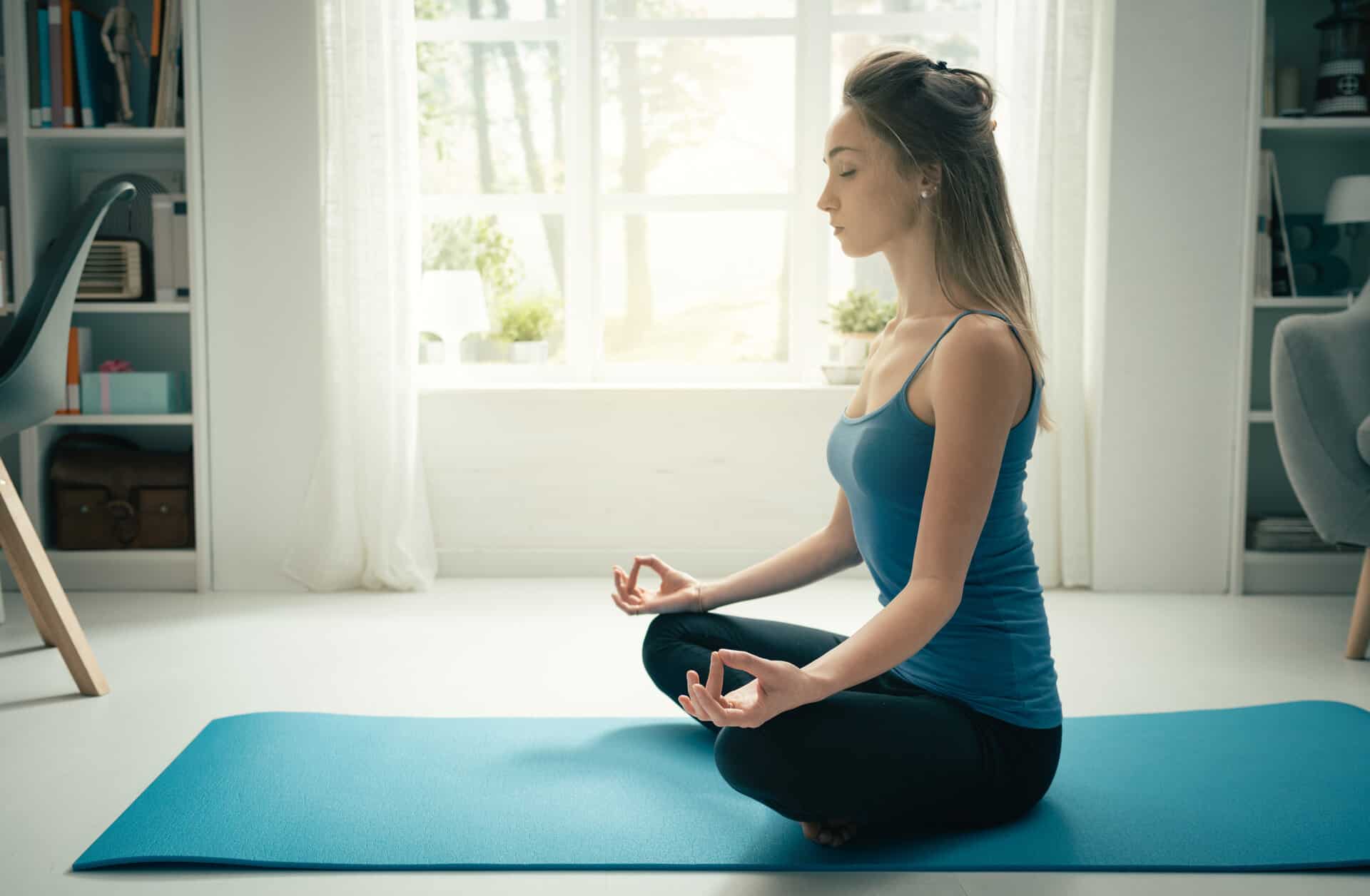 Young confident woman practicing mindfulness meditation at home, she is sitting with crossed legs on a mat