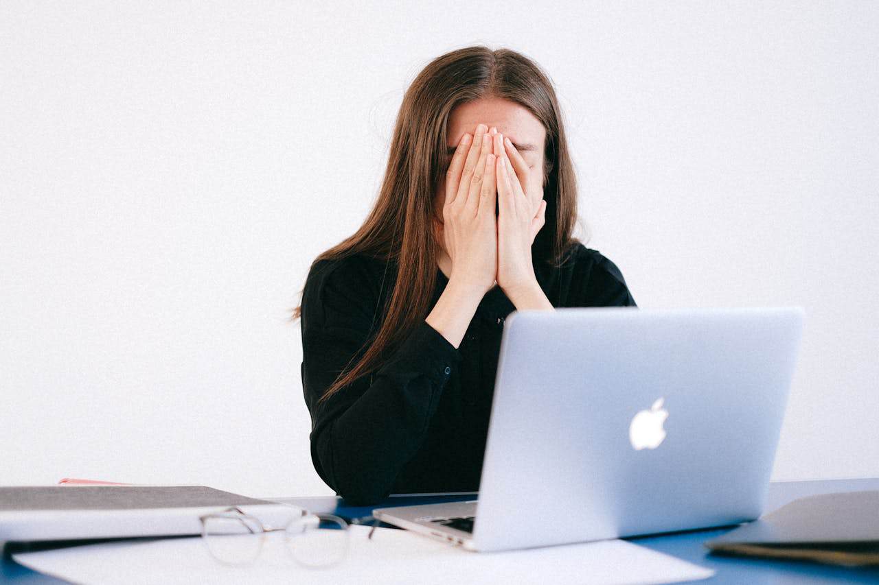 A stressed young woman wearing a black shirt sitting at a desk with an open laptop, covering her face with both hands.