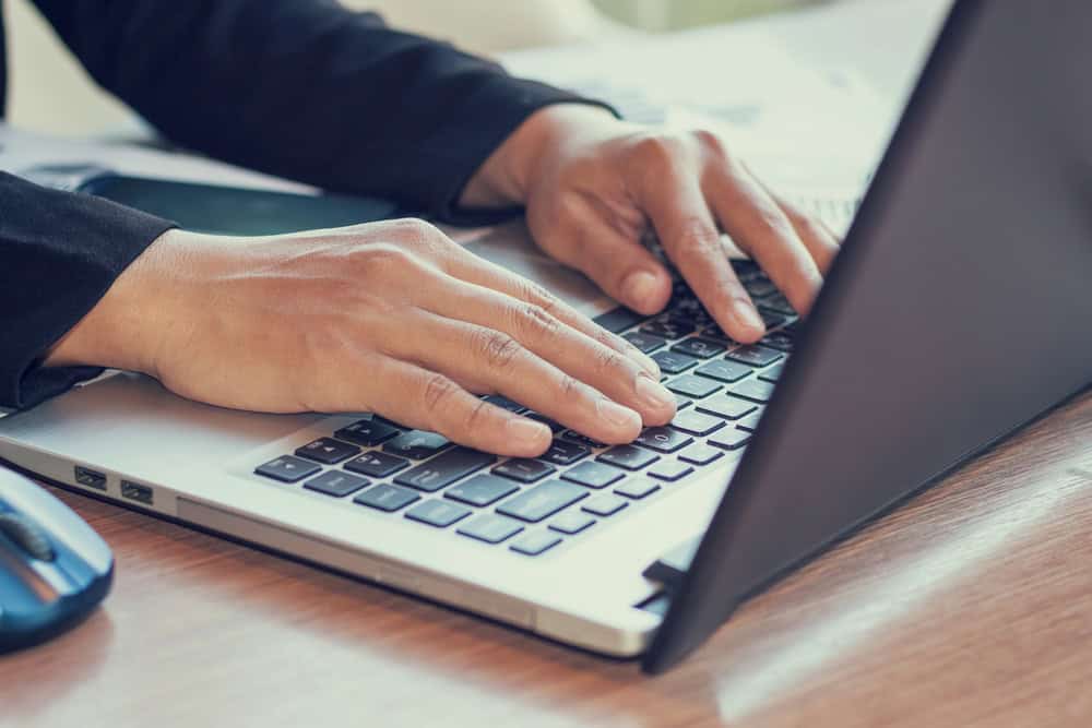 A close-up of a person's hands wearing a dark suit jacket typing on a silver laptop on a wooden desk.