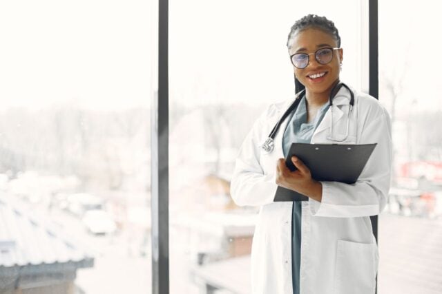A smiling Black female doctor wearing glasses, a white lab coat, and a stethoscope, holding a clipboard while standing next to a bright window